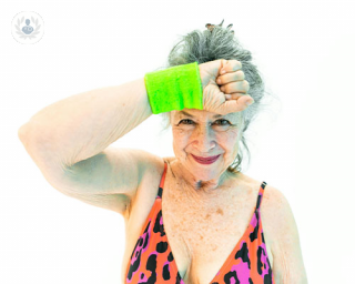 An older woman grins at the camera in loud, 80s workout gear. She wipes a sweatband over hear forhead