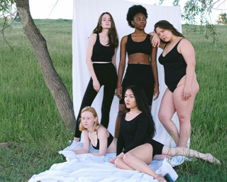 A group of women standing by a backdrop set up outside on the grass beneath an old tree. They are multiracial and have different skin, hair, and body types. They are all wearing black shorts and shirts to best highlight their natural diversity.