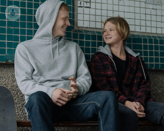 Two teenage boys sitting together against a wall and smiling at each other