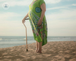 A woman stands on the shore at sunset and looks pensively to the horizon. She holds a walking cane but she isn't reliant on it.