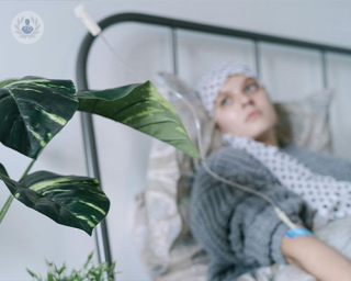 A young woman wearing a head scarf and with an IV reclines in bed and looks wistfully out of a window