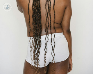 A person stands with their back to the camera. They are shirtless and wearing white briefs. They have long black braids that travel down their back.