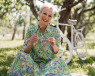 A smiling elderly woman wearing a green dress. She is sat on the grass, holding her sunglasses off her face